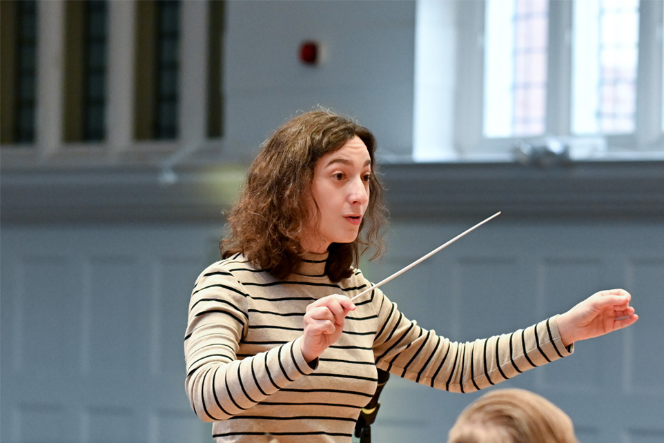 Postgraduate student Michal Oren, a female student, with medium height brown hair, wearing a white and black striped shirt, conducting a small orchestra in the Amaryllis Fleming Concert Hall.